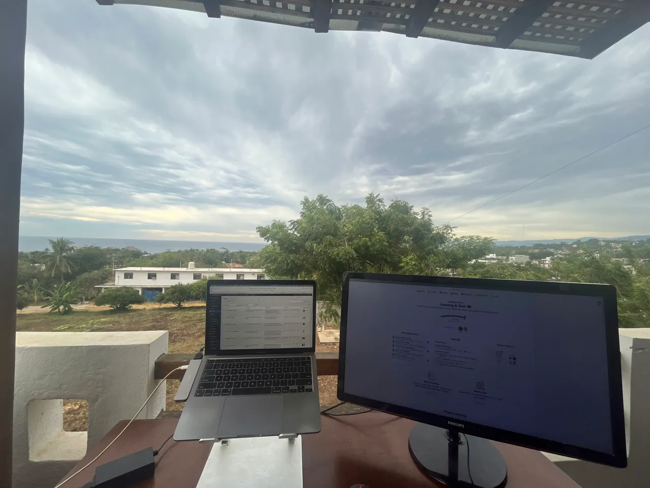 Patrick's outdoor workspace with a laptop and a monitor, set against a green landscape and a cloudy sky.